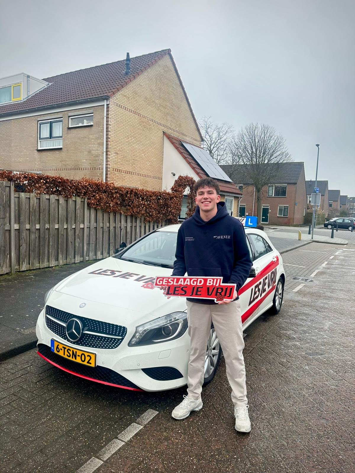 Geslaagde leerling uit het Westland met het bord Geslaagd naast de lesauto van Rijschool Les je Vrij
